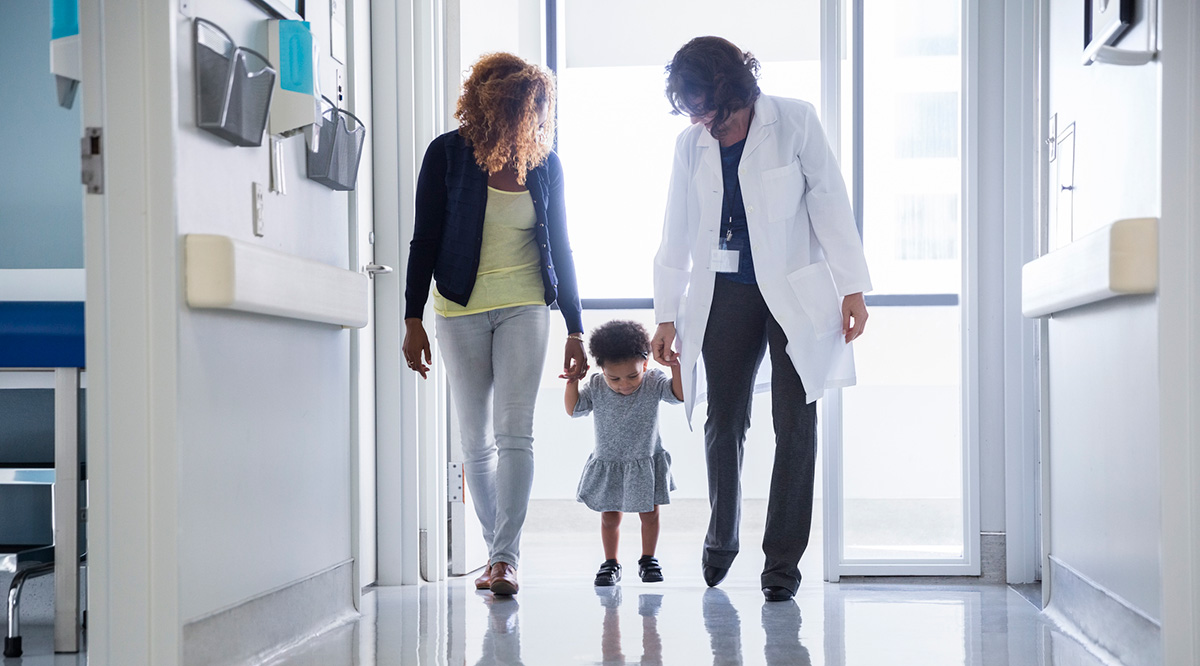 Mother and pediatrician holding hands with toddler while walking in hospital corridor